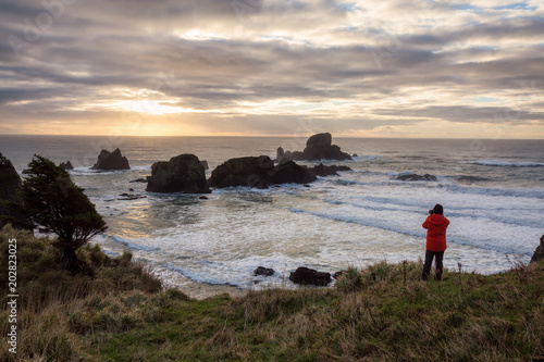 Man with a camera is looking at a Beautiful Pacific Ocean Coast with rugged rocky formation in the background. Taken in Ecola State Park, Seaside, Cannon Beach, Oregon, USA.