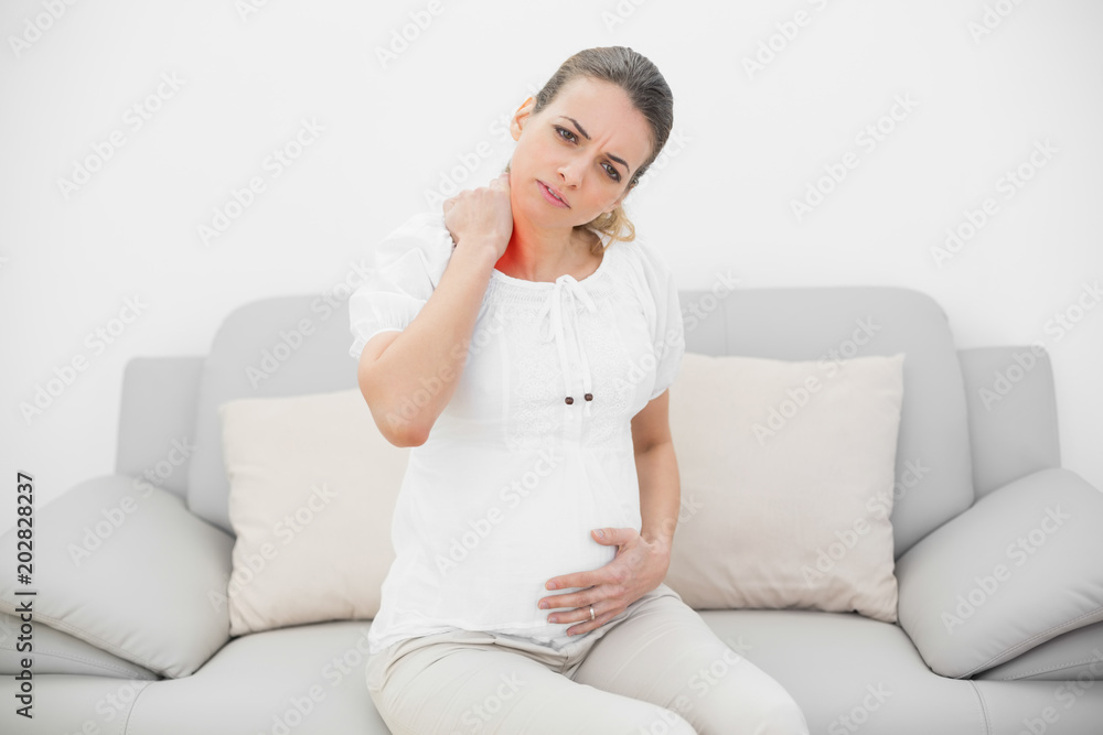 Attractive pregnant woman holding her injured neck while touching her belly and sitting on couch Attractive pregnant woman holding her injured neck while touching her belly and sitting on couch