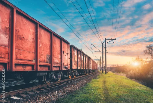 Naklejka na ścianę Czerwoni ładunków furgony i pociąg na staci kolejowej przeciw niebieskiemu niebu z chmurami przy zmierzchem. Kolorowy przemysłowy krajobraz z linią kolejową. Peron. Przemysł ciężki. Wysyłka ładunków. Transport