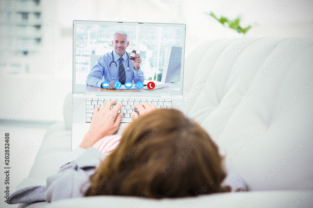 Doctor holding medicine jar against woman using laptop while lying on sofa Doctor holding medicine jar against woman using laptop while lying on sofa