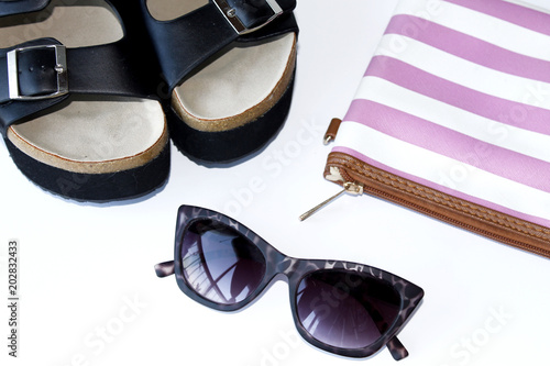 Sandalias de tiras negras de verano, gafas de sol y cartera de mano a rayas blanca y rosa. Fondo blanco