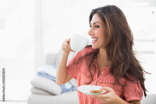 Happy brunette sitting on her sofa holding cup and saucer with a cookie