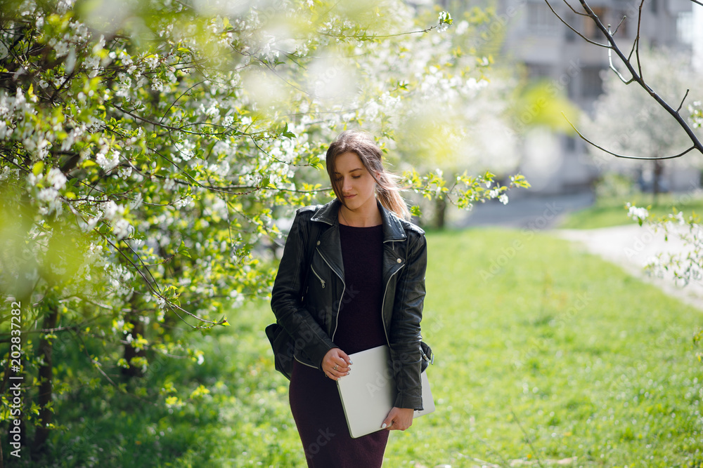 Young handsome brunette woman in black leather jacket holding white laptop computer while standing in the park near green trees Young handsome brunette woman in black leather jacket holding white laptop computer while standing in the park near green trees