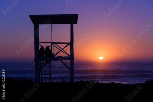 Romantic scene of couple silhouette on lifeguard tower watching splendid sunset on empty beach in La Serena, Chile. Summertime, travel destination, Valentines Day concept