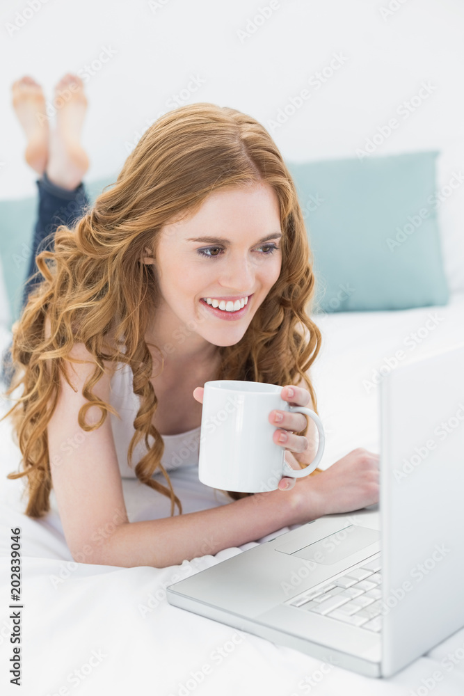 Smiling blond with coffee cup using laptop in bed Smiling blond with coffee cup using laptop in bed