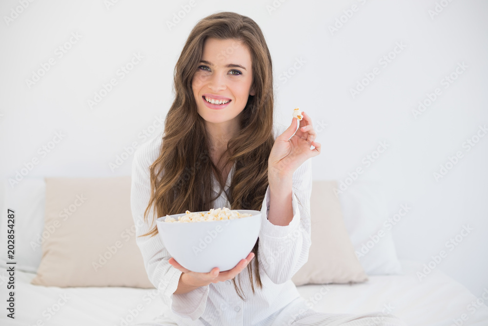 Delighted casual brown haired woman in white pajamas eating popcorn Delighted casual brown haired woman in white pajamas eating popcorn
