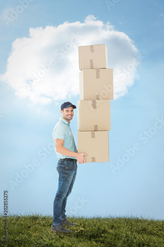 Confident delivery man carrying stack of boxes against blue sky over green field