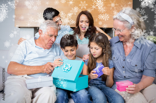 Extended family sitting on sofa with gift boxes in living room against snowflakes