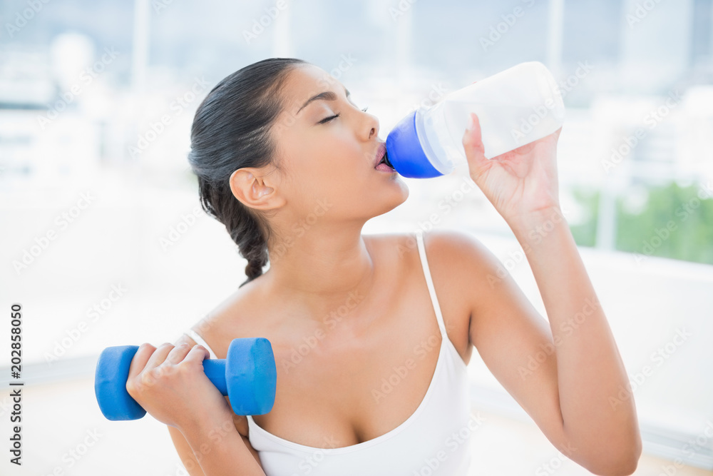 Peaceful toned brunette with dumbbells drinking from sports bottle Peaceful toned brunette with dumbbells drinking from sports bottle