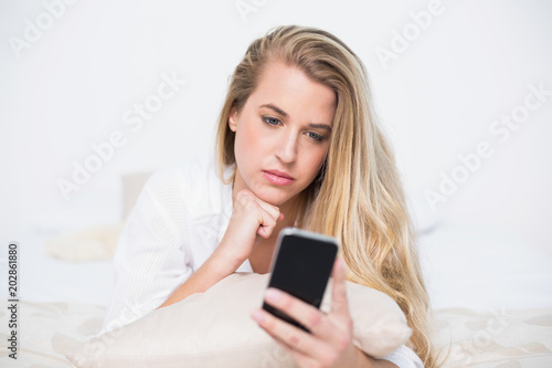 Pensive gorgeous model looking at her smartphone lying on cosy bed