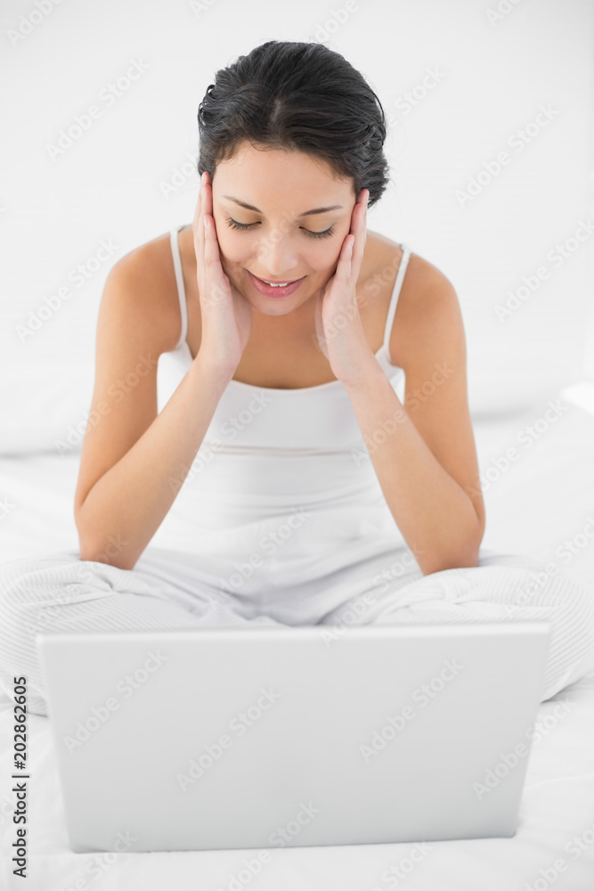 Dreamy casual brunette in white pajamas sitting on her bed and looking at her laptop Dreamy casual brunette in white pajamas sitting on her bed and looking at her laptop