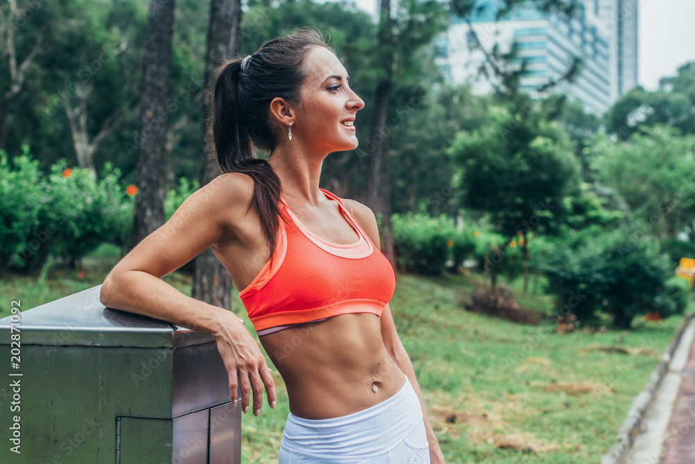 Slim fitness brunette woman with six pack abs wearing pink sport bra  standing in city park relaxing after workout looking away from the camera  Stock Photo