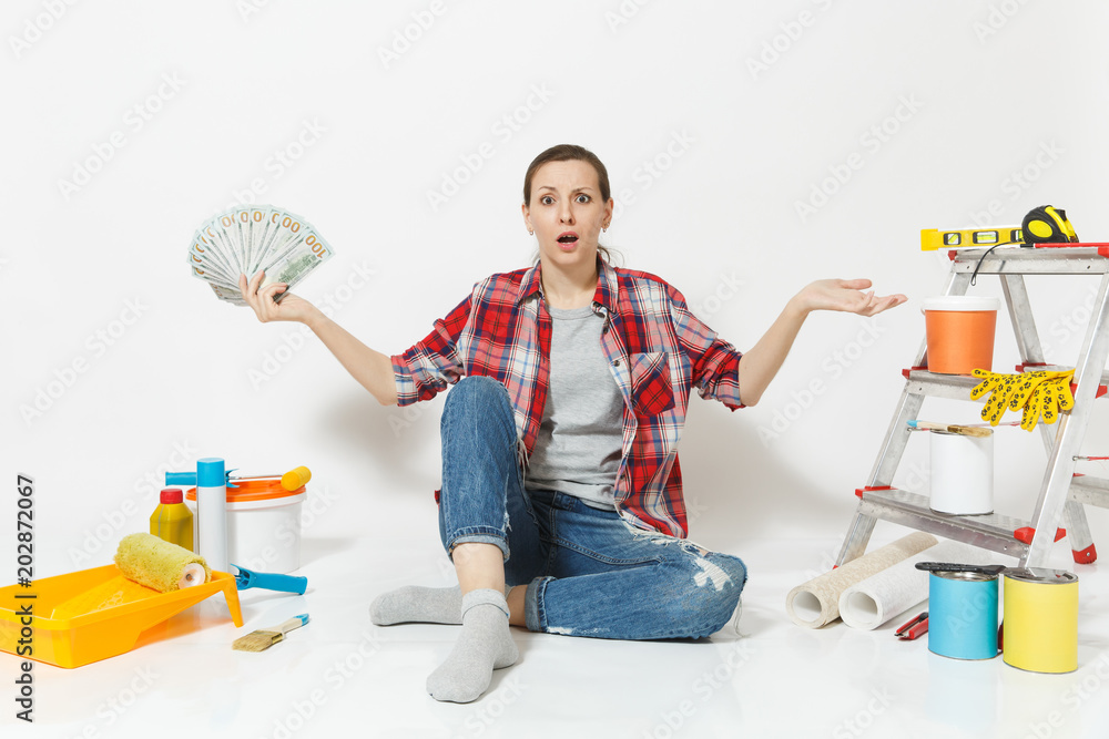 Woman in shock holds bundle of dollars, cash money, sits on floor with instruments for renovation apartment isolated on white background. Wallpaper accessories for gluing tools. Concept of repair home Woman in shock holds bundle of dollars, cash money, sits on floor with instruments for renovation apartment isolated on white background. Wallpaper accessories for gluing tools. Concept of repair home