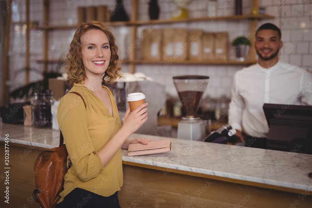 Portrait of smiling young female customer holding disposable coffee cup at counter against waiter