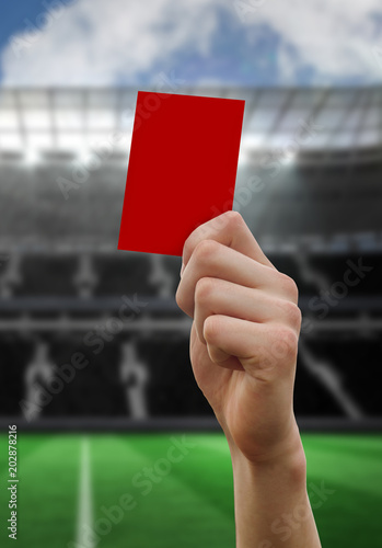 Hand holding up red card against large football stadium with empty stands