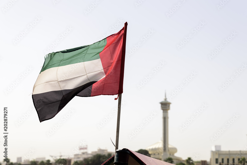 Obraz premium Powiewająca na wietrze flaga ZEA na zatoce Dubai Creek ze starą dzielnicą Dubaju, Bur Dubai i minaretem meczetu w tle