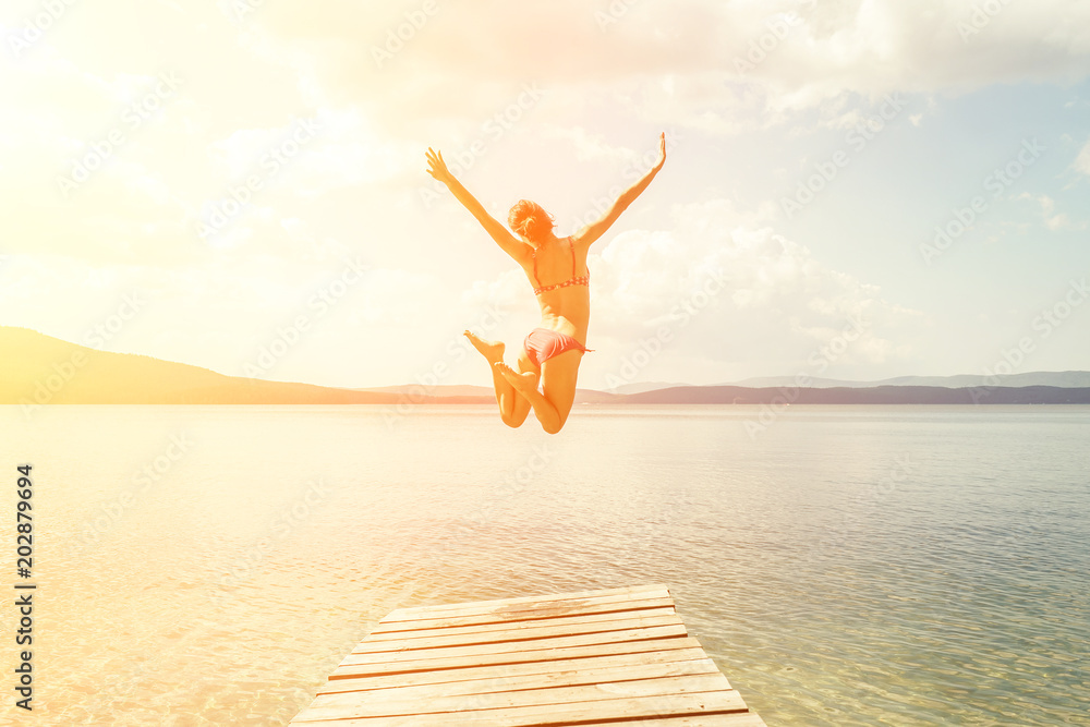 The girl in a swimsuit is jumping from a wooden pier into the water against the blue mountains. On the shore of the ocean, the sea, the lake. Happiness, summer, fun