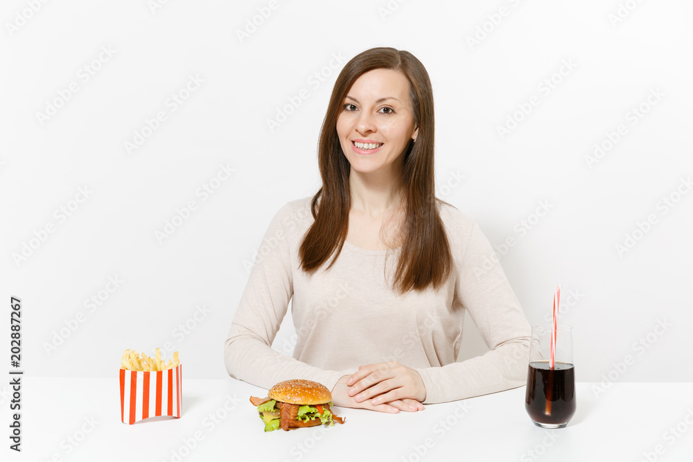 Beautiful smiling woman sitting at table with burger, french fries, cola in glass bottle isolated on white background. Proper nutrition or American classic fast food. Advertising area with copy space. Beautiful smiling woman sitting at table with burger, french fries, cola in glass bottle isolated on white background. Proper nutrition or American classic fast food. Advertising area with copy space.