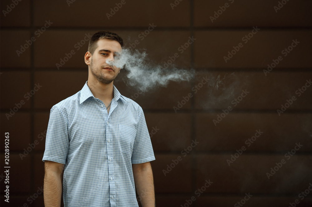 Vape man. Portrait of a handsome young white guy with modern haircut in in checkered sleeveless shirt vaping an electronic cigarette opposite dark background in the spring. Lifestyle. Vape man. Portrait of a handsome young white guy with modern haircut in in checkered sleeveless shirt vaping an electronic cigarette opposite dark background in the spring. Lifestyle.