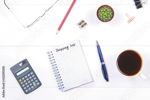 Notepad with the text: a shopping list. White table with calculator, cactus, note paper, coffee mug, pen, glasses.