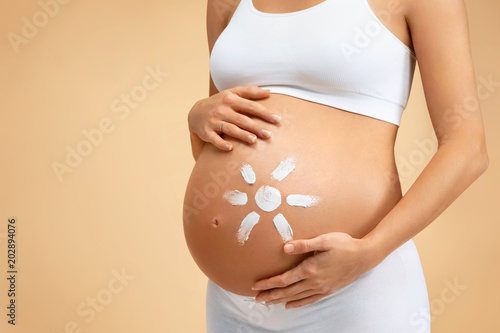 Close up of pregnant woman applying moisturizing cream on her belly on beige background. Pregnancy, maternity, preparation and expectation concept