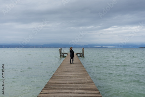 Woman standing in the middle of a pier on the Lake di Garda, italy, with snow capped mountains and clouds in the background