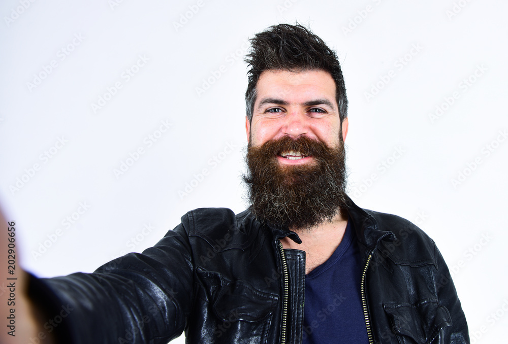Man with beard and mustache on smiling face looking at camera. Macho wears leather jacket, white background. Hipster looks happy and cheerful while taking selfie photo. Masculinity concept. Man with beard and mustache on smiling face looking at camera. Macho wears leather jacket, white background. Hipster looks happy and cheerful while taking selfie photo. Masculinity concept.