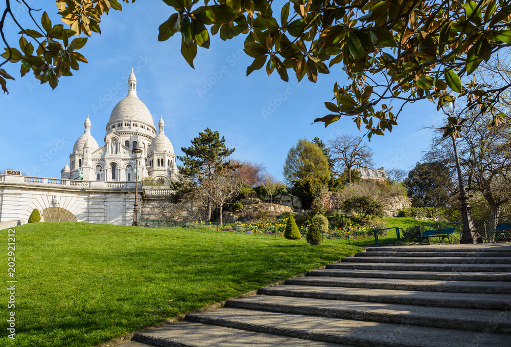 The bright white basilica of the Sacred Heart of Paris situated at the top of the Montmartre hill, seen from the Louise Michel park by a sunny spring morning with foliage and stairs in the foreground.