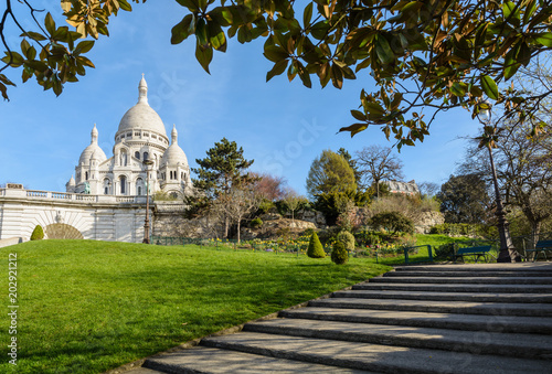 The bright white basilica of the Sacred Heart of Paris situated at the top of the Montmartre hill, seen from the Louise Michel park by a sunny spring morning with foliage and stairs in the foreground.