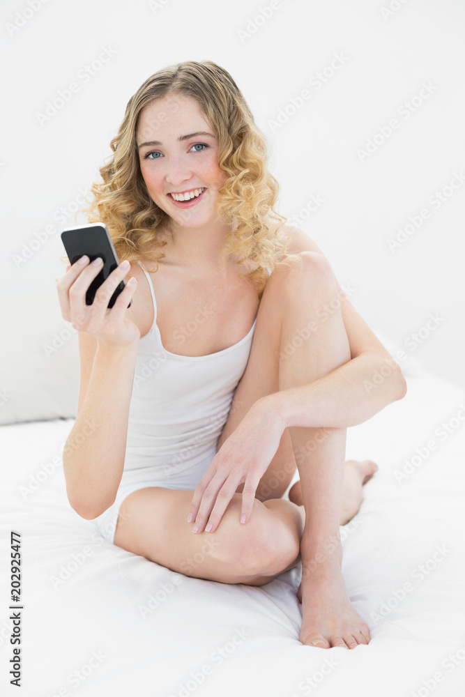 Pretty smiling blonde sitting on bed holding smartphone Pretty smiling blonde sitting on bed holding smartphone