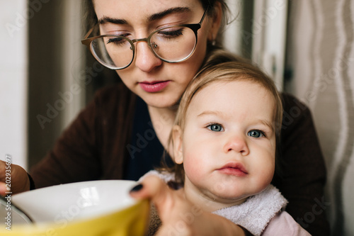 Young happy mother feeding her little baby with delicious tasty soup from plate in kitechen. Funny portrait of eating child. Beautiful girl in glasses together with daughter have diinner at home.
