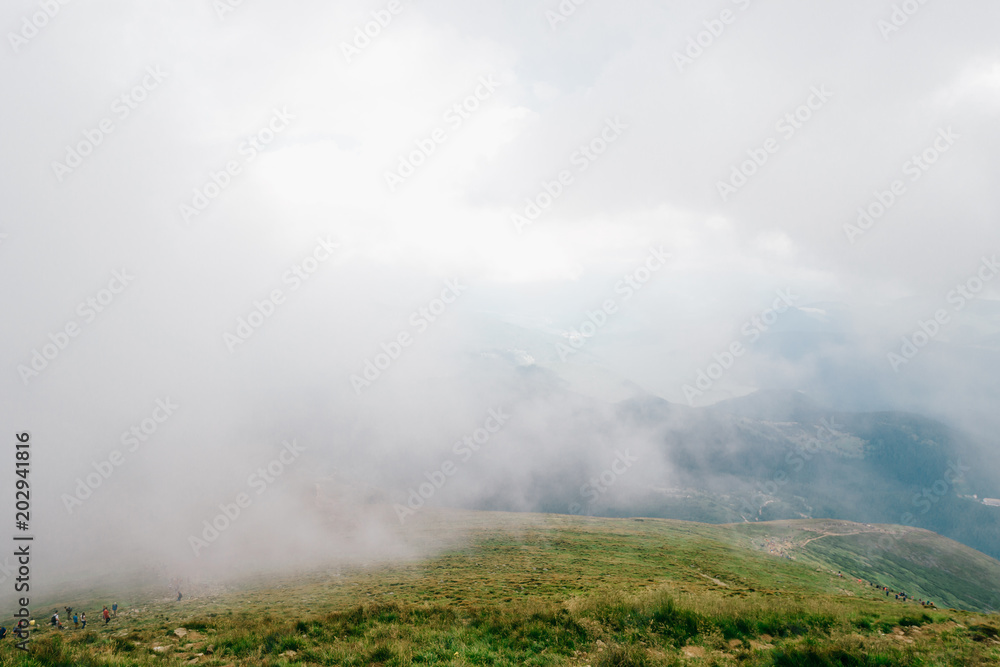 Foggy weather in mountains. Travelers hiking over misty hiils. Tourists climbing up. Healthy  activity. Summer moody scenic view from high altitude at nature outdoor. Beauftiful landscape. Trekking.
