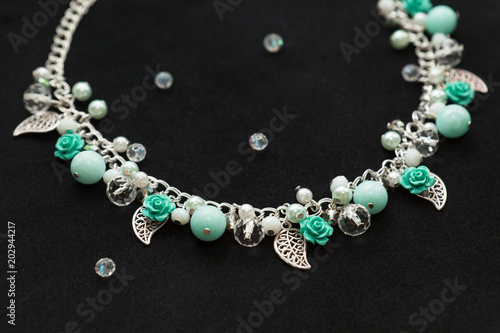 Beautiful handmade turquoise necklace with shiny crystals, gemstone beads, plastic flowers and metallic leaves, lying on the black background, side perspective view