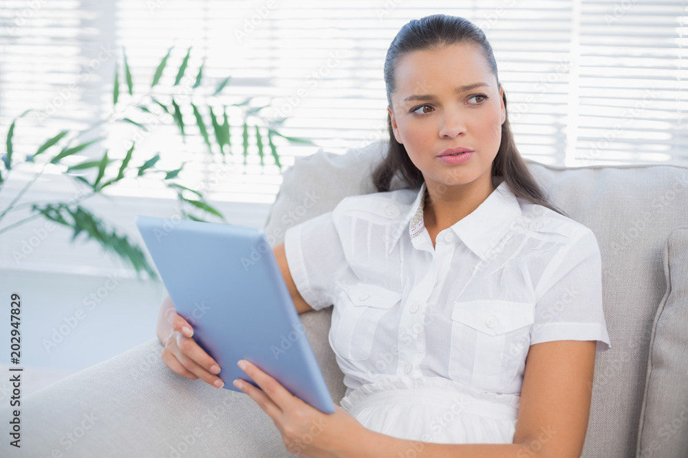 Thoughtful cute woman using tablet sitting on cosy sofa Thoughtful cute woman using tablet sitting on cosy sofa