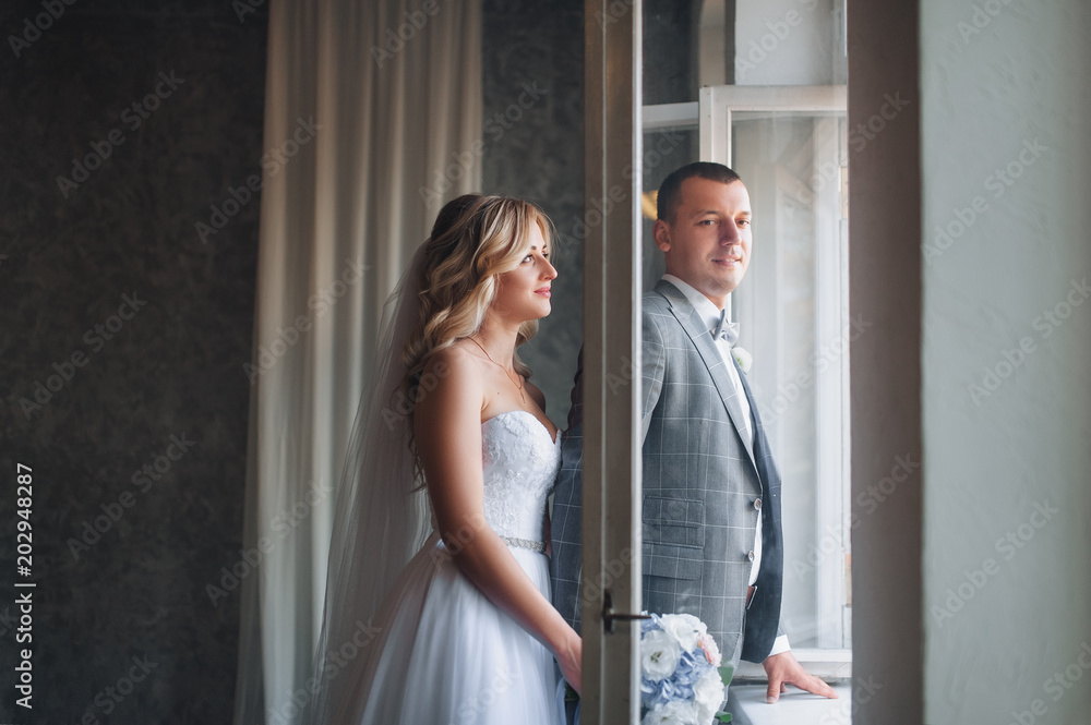 Beautiful bride and groom with curly hair stand near the window in the studio. Portrait of stylish newlyweds. Beautiful bride and groom with curly hair stand near the window in the studio. Portrait of stylish newlyweds.