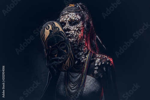 Make-up concept. Portrait of a scary African shaman female with a petrified cracked skin and dreadlocks, holds a traditional mask on a dark background. Make-up concept. photo