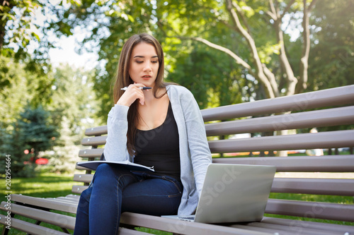 Young female IT student on a bench,. Doing homework in the park with laptop and textbook