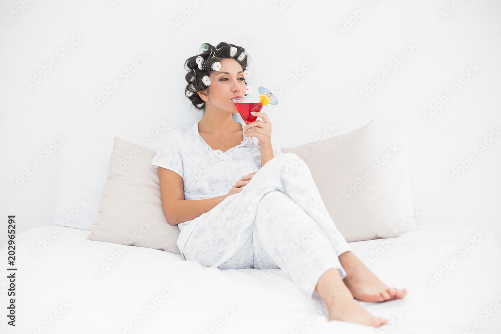 Smiling brunette in hair curlers sitting on her bed drinking a cocktail Smiling brunette in hair curlers sitting on her bed drinking a cocktail