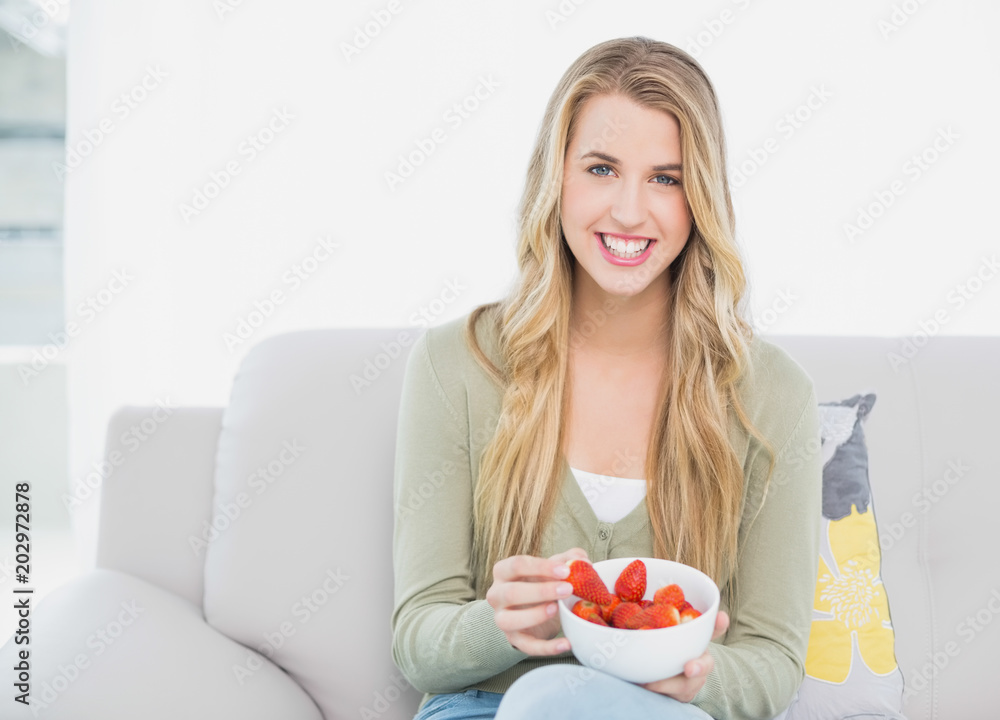 Smiling pretty blonde eating strawberries sitting on cosy sofa Smiling pretty blonde eating strawberries sitting on cosy sofa