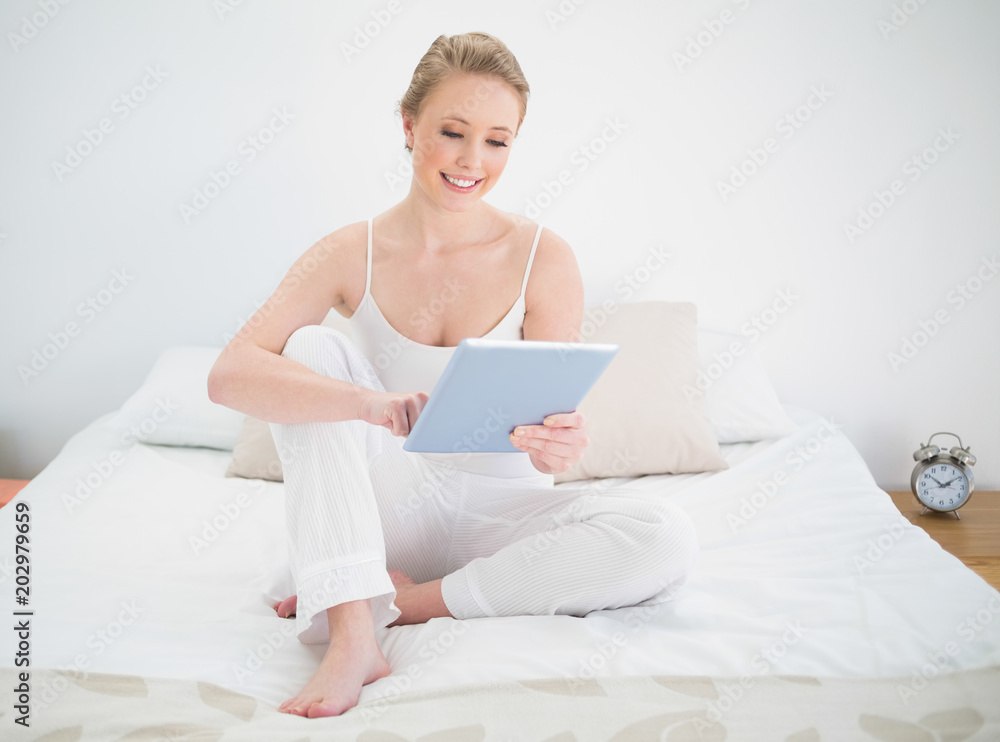 Natural smiling blonde using tablet while sitting on bed Natural smiling blonde using tablet while sitting on bed