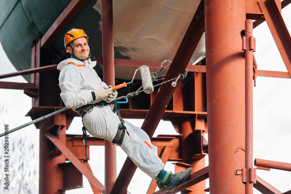 Industrial climber in helmet and uniform painting water tower. Professional Painter working on height. Risky job. Extreme occupation. Industrial climber in helmet and uniform painting water tower. Professional Painter working on height. Risky job. Extreme occupation.