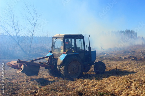 Blue tractor rides through the burnt field against a blue smoke to plow a protective strip of forest
