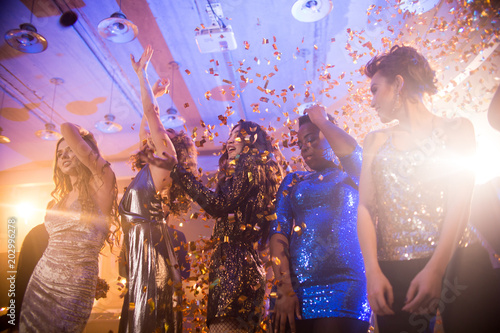 Group of beautiful young women wearing glittering dresses dancing under golden confetti shower enjoying party in nightclub