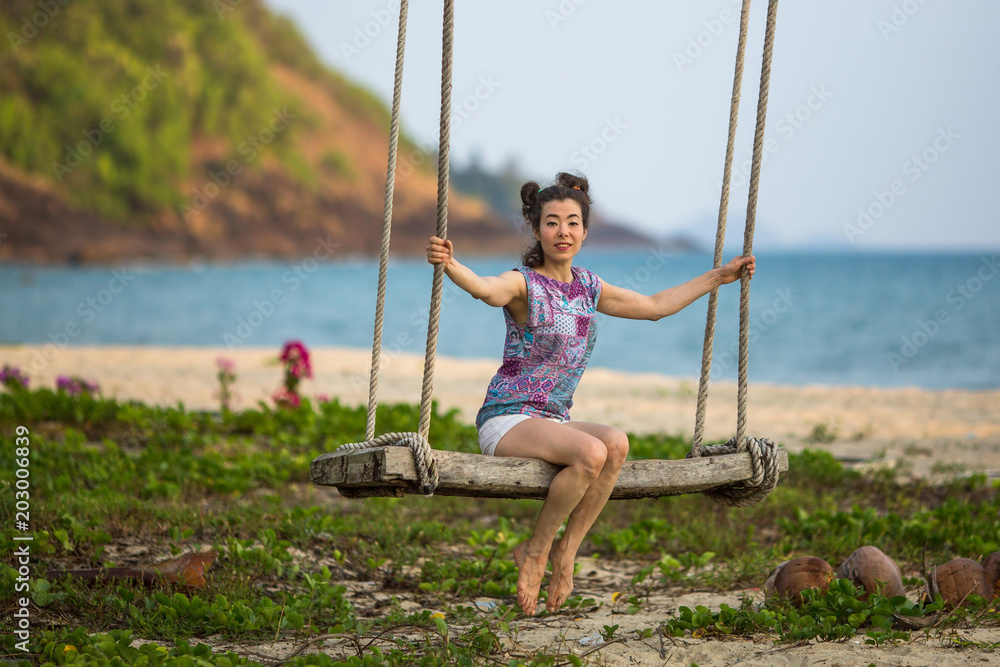 A young mixed race woman swinging on a wooden swing at a seaside tropical beach.