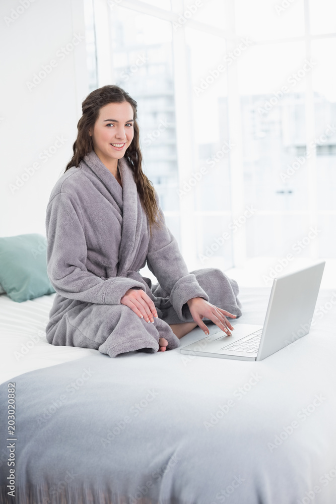 Smiling brunette in bathrobe using laptop in bed Smiling brunette in bathrobe using laptop in bed