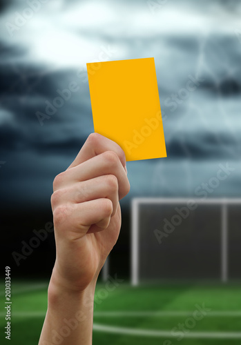 Hand holding up yellow card against football pitch under stormy sky
