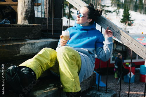 Young woman snowboarder wearing sunglasses drinking coffee from glass cap outdoor in Winter Ski resort