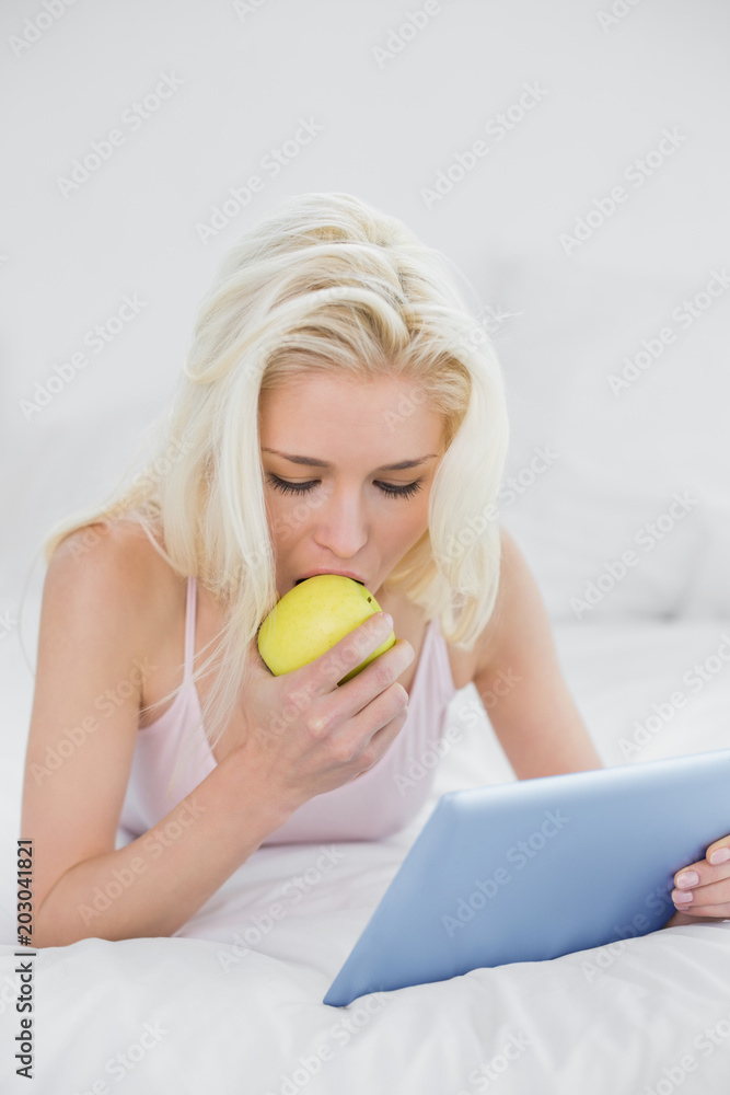 Casual blond eating an apple while using tablet PC in bed Casual blond eating an apple while using tablet PC in bed