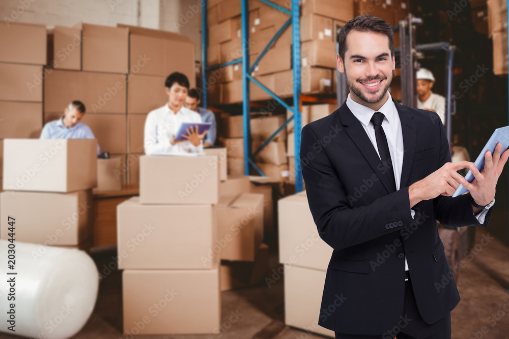Businessman using his tablet while looking at the camera against people at work in warehouse Businessman using his tablet while looking at the camera against people at work in warehouse