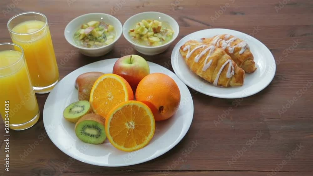 On a wooden table are a plate of fruit and two glasses of orange juice. The camera moves from left to right. On a wooden table are a plate of fruit and two glasses of orange juice. The camera moves from left to right.